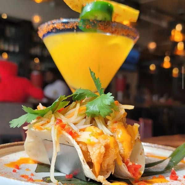 Krave's Jamaican tacos with slaw and sauce, paired with a vibrant yellow cocktail garnished with jalapeño.