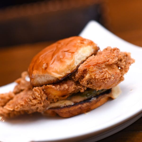 Crispy fried chicken sandwich with pickles and sauce on a toasted bun, served on a white plate.